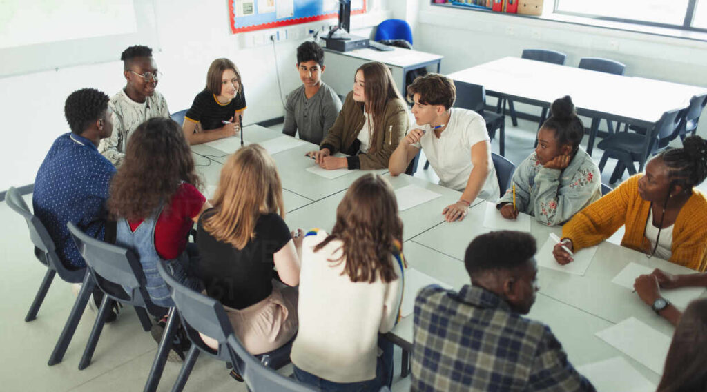 A group of students sit around a table having a discussion in a classroom.