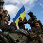 Men stand and sit on a tank, a Ukrainian flag waves in the background.