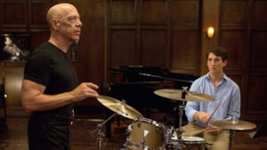 Two men sit and stand around a drum set.