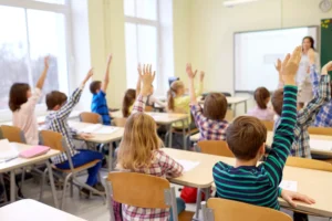 Kids raising hands in a classroom with a teacher gesticulating.