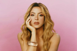 Shakira poses in front of a pink background with her hand on her face.